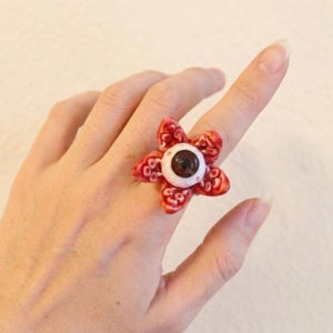 May include: A unique ring featuring a red, flower-shaped design with a central brown eye. The petals have a textured appearance, and the ring is worn on a finger. The ring's design is reminiscent of a Demogorgon.