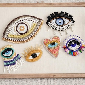 Könnte beinhalten: Eine Sammlung handgefertigter, augenförmiger Broschen. Die Broschen weisen aufwendige Perlenarbeiten in verschiedenen Farben auf, darunter Braun, Gold, Schwarz, Blau und Rosa. Einige haben Perlen- und Kristallakzente, und eine ist herzförmig.