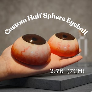 以下が含まれることがあります： 茶色の虹彩と詳細な赤い静脈を持つ、2つのカスタムハーフスフィア眼球。テキスト「Custom Half Sphere Eyeball」は眼球の上にアーチ状に配置されています。サイズは7cmです。