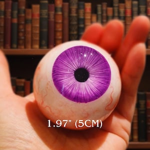 Op de afbeelding: Een realistische, bolvormige oogbal met een levendige paarse iris en een zwarte pupil. Het oog heeft een diameter van 5 cm, met zichtbare rode aderen. De achtergrond toont een vage boekenkast.