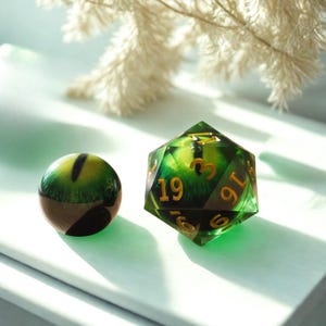May include: A green and gold D20 die and a round eye-shaped object. The D20 has gold numbering and a green interior with a textured pattern. The round object has a green and yellow eye design.