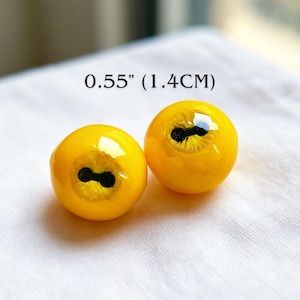 Op de afbeelding: Twee ronde, felgele cabochons met een gladde, glanzende afwerking. Elk stuk heeft een centrale zwarte, horizontale pupilvorm omringd door een textuurrijke, glinsterende iris. De tekst 1.4CM staat erboven om de afmeting aan te geven.