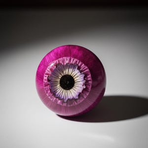 May include: A spherical, magenta decorative item with a floral pattern. The centre has a black circle encircled by white and purple petals. The object is smooth and glossy, casting a shadow on a white surface.