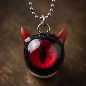 May include: A pendant necklace featuring a black and red devil eye design. The pendant has a black base with a large red eye and red horns. The necklace chain is a silver ball chain.