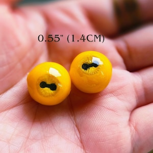 May include: Two round, bright yellow cabochons with a smooth, glossy finish and a central black, horizontal pupil design. The text 0.55" (1.4CM) is visible above the objects, indicating their diameter. These items have a domed, eye-like appearance.