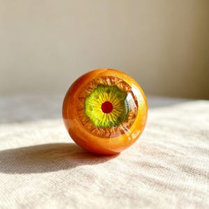 May include: A spherical object with a glossy orange exterior featuring a central iris design. The iris is composed of a vibrant green ring surrounding a small, solid red pupil, creating a realistic eye-like appearance with a smooth, reflective finish.