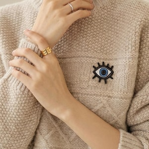May include: A beige cable-knit sweater with a decorative eye-shaped brooch. The brooch features a white iris, a blue pupil, and black beaded details. A gold ring and a silver ring are also visible.