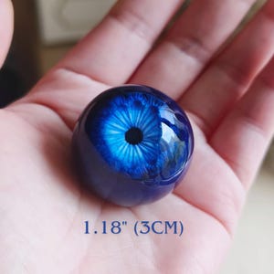 May include: A spherical glass eye cabochon featuring a deep blue iris with intricate radial patterns and a solid black pupil. The object has a smooth, glossy finish and measures 1.18 inches in diameter, providing a realistic appearance for crafts.