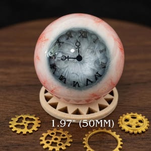 Puede incluir: Un objeto decorativo con forma de globo ocular con un diseño de esfera de reloj, que mide 50 mm de diámetro. El globo ocular tiene venas rojas y blancas, una esfera de reloj gris y está colocado sobre un soporte de madera. Engranajes dorados están esparcidos alrededor de la base.