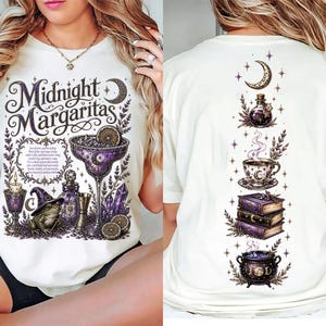 May include: A white t-shirt featuring a front graphic with the text Midnight Margaritas, a margarita glass, a frog in a witch hat, a candle, and crystals. The back graphic displays a vertical arrangement of a crescent moon, potion, teacup, books, and cauldron.