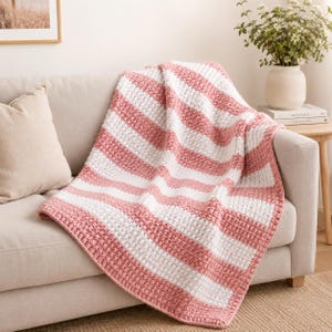 May include: A pink and white striped crochet blanket draped over a beige sofa. The blanket features thick, alternating stripes of white and a soft pink. The texture of the blanket is visible, suggesting a handmade item. The sofa has a beige pillow.
