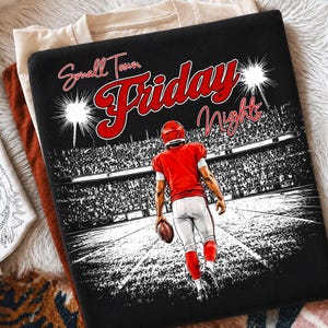 May include: A black t-shirt featuring a graphic of a football player in a red jersey and white pants holding a football on a field. The text "Small Town Friday Nights" is printed in red and white script above the stadium crowd scene.