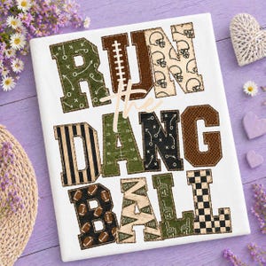 May include: A white rectangular sign featuring the text RUN THE DANG BALL in large block letters. The letters are filled with various football-themed patterns including helmets, play diagrams, stripes, footballs, and checkered designs.