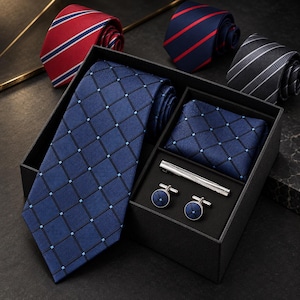 May include: A blue necktie with a black grid and light blue dot pattern, a matching pocket square, a silver tie bar, and two round blue cufflinks in a black gift box. Three additional rolled neckties in red, navy, and grey stripes are nearby.