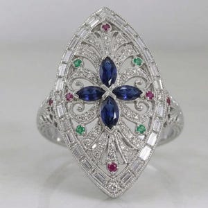 Puede incluir: Un anillo plateado con un diseño ovalado alargado y ornamentado. El anillo presenta un arreglo floral central de piedras preciosas azules, rodeado de piedras rojas y verdes más pequeñas y piedras claras con corte baguette. La banda tiene un patrón de filigrana detallado.
