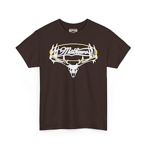 May include: A brown t-shirt featuring a white graphic of a deer skull with antlers and the name "Mathews" in a stylized font, enclosed in a yellow oval. The shirt has a crew neck and short sleeves.