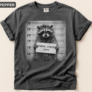 May include: A charcoal gray crew neck t-shirt featuring a black and white graphic of a raccoon in a mugshot pose. The raccoon holds a sign that reads FERAL SINCE 2005 against a height chart background with imperial measurements in feet and inches.