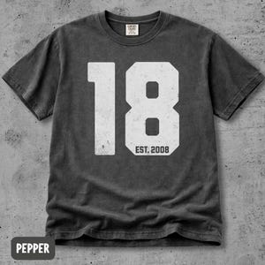 Peut inclure: Un t-shirt à manches courtes gris anthracite arborant un grand graphisme blanc vieilli du chiffre 18 avec le texte EST. 2008 centré sous le nombre. Le t-shirt présente une encolure ronde et une étiquette de marque Comfort Colors au col.