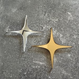 May include: Two four-pointed star ornaments. One is silver, with a shiny, reflective surface. The other is gold, with a matte finish. Both are placed on a gray, textured surface. These decorative stars could be used for crafts or home decor.