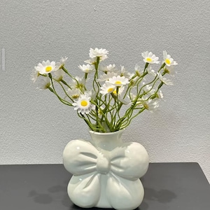 Pode incluir: Um vaso de cerâmica branco em forma de laço, cheio de flores de margarida brancas com centros amarelos. O vaso está sobre uma superfície cinza escuro contra uma parede cinza claro. As flores têm caules verdes.