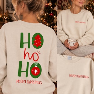 Pode incluir: Uma camisola de gola redonda bege com um gr&aacute;fico vermelho e verde nas costas com o texto HO HO HO, uma &aacute;rvore de Natal vermelha e um enfeite vermelho. A parte frontal do peito apresenta o texto vermelho MERRY CHRISTMAS em fonte simples.