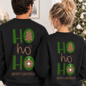 Pode incluir: Duas camisolas pretas de gola redonda com uma impress&atilde;o gr&aacute;fica nas costas. O design apresenta o texto HO HO HO em verde e castanho, com uma &aacute;rvore de Natal e um enfeite castanho a substituir as letras O. O texto MERRY CHRISTMAS est&aacute; abaixo.