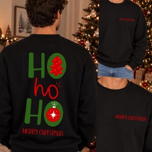 Pode incluir: Uma camisola preta de manga comprida com um gr&aacute;fico nas costas que inclui o texto verde HO HO HO, uma &aacute;rvore de Natal vermelha, um ornamento vermelho e o texto vermelho MERRY CHRISTMAS. O peito exibe o texto vermelho MERRY CHRISTMAS.