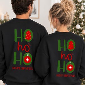 Pode incluir: Duas camisolas pretas de gola redonda com um gr&aacute;fico nas costas apresentando o texto HO ho HO em verde e vermelho. O design inclui uma &aacute;rvore de Natal vermelha dentro de um oval verde e um enfeite de Natal vermelho num oval verde, com Merry Christmas em vermelho.