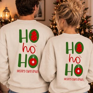 Pode incluir: Duas camisolas bege de gola redonda com um gr&aacute;fico nas costas apresentando o texto verde HO ho HO. O primeiro O cont&eacute;m uma &aacute;rvore de Natal vermelha e o segundo O uma bola de Natal vermelha. O texto vermelho MERRY CHRISTMAS est&aacute; abaixo.