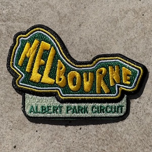 Puede incluir: Parche bordado con la palabra "MELBOURNE" en amarillo, contorneada en verde y negro. El texto "ALBERT PARK CIRCUIT" está debajo. El parche tiene una forma única y es probablemente para una chaqueta o bolso.