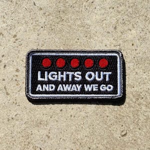 May include: A black rectangular patch with white embroidered text: "LIGHTS OUT AND AWAY WE GO". The patch features four red circles above the text and a black and white stitched border. The patch is designed to be sewn on.