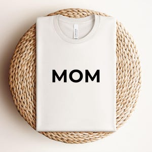 May include: A white ceramic mug with a black handle and the text "But First, Coffee" in a black, handwritten-style font. The mug is on a white surface.