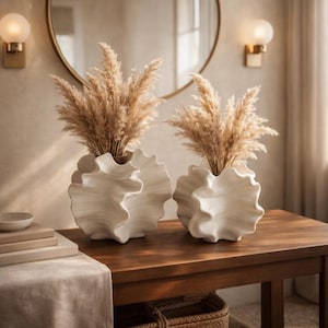 May include: Two white, ruffled ceramic vases filled with dried pampas grass sit on a wooden table. The vases have a unique, wave-like design. The table is made of dark wood and has a woven basket underneath.