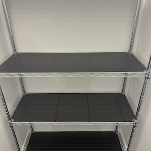 May include: A chrome wire shelving unit with four shelves. Each shelf has a black, textured liner. The unit is empty and positioned against a white wall. The shelves are evenly spaced.