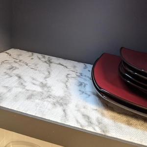 May include: A stack of four burgundy-colored, oval-shaped plates with a dark rim. The plates are on a shelf lined with a white and gray marble-patterned shelf liner. The shelf is inside a dark gray cabinet.