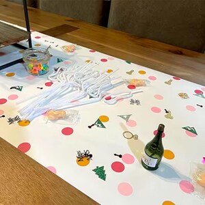 May include: A festive party table setting with a white tablecloth decorated with pink and orange polka dots, and confetti. Includes a small green bottle, clear straws, and a jar of colorful candies. The table is set for a celebration.