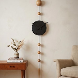 Könnte beinhalten: Moderne Wanduhr mit schwarzem, kreisförmigem Zifferblatt und schwarzen Zeigern. Die Uhr hängt an einer schwarzen Stange mit Holzkugeln und einem tropfenförmigen Holzelement am unteren Ende. Die Uhr ist an einer cremefarbenen Wand befestigt.