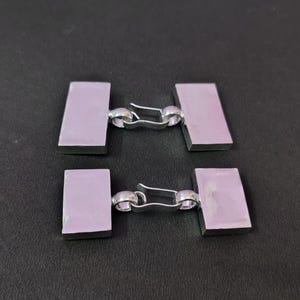 May include: A pair of rectangular cufflinks featuring a smooth, light lavender-colored surface set within a polished silver-toned metal frame. Each cufflink consists of two rectangular plates connected by a curved, hinged silver-toned metal bar.