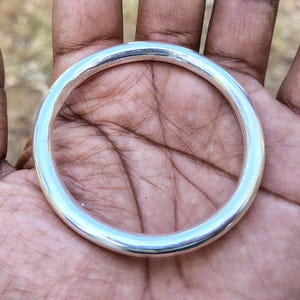 May include: A smooth, polished silver-colored circular bangle bracelet with a rounded profile. The metal surface is reflective and uniform in thickness, forming a simple, continuous loop without any visible clasps, hinges, or decorative engravings.