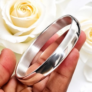 May include: A polished silver-toned bangle bracelet with a smooth, rounded exterior surface and a brushed metallic finish on the interior. The circular band features a reflective, high-shine appearance that catches light across its curved profile.