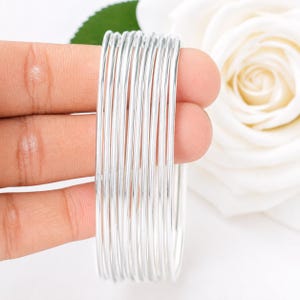 Peut inclure: Un ensemble de dix bracelets joncs fins et polis de couleur argentée, disposés en pile parallèle. Chaque bracelet individuel présente une surface lisse et arrondie avec une finition métallique réfléchissante, créant une apparence uniforme et cohérente.