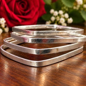 May include: A set of three stacked silver-toned bangles with a square shape and a smooth, reflective metallic finish. The bracelets are arranged in a tiered formation, showcasing their uniform thickness and rounded corners.