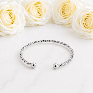 May include: A silver-toned metal cuff bracelet featuring a twisted rope-like texture throughout the band. The bracelet has an open design with a smooth, polished spherical ball at each end, creating a clean and reflective metallic appearance.