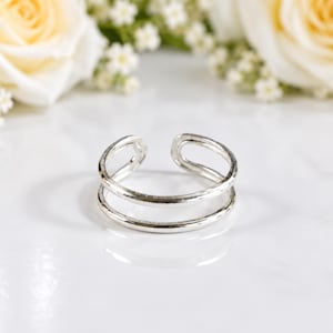 Könnte beinhalten: Ein silberfarbener offener Ring mit einem Doppelband-Design und einer glatten, polierten Oberfläche. Der Ring weist oben eine Lücke auf, wodurch zwei abgerundete, parallele Schlaufen entstehen, die an der Basis verbunden sind und eine durchgehende, glatte und reflektierende Metallstruktur bilden.