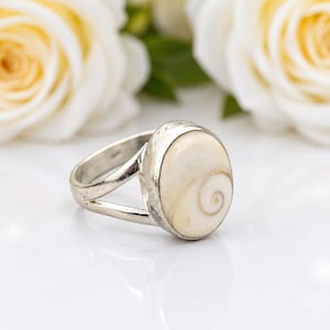 Puede incluir: Un anillo de plata con una concha de ojo de Shiva ovalada y lisa como pieza central. La banda está dividida en dos secciones distintas que se unen en el engaste, el cual sostiene la concha de color crema con su patrón de espiral marrón.
