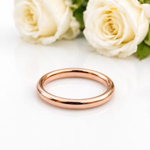 Op de afbeelding: Een gepolijste ring van roségoud met een glad, afgerond oppervlak. De ring is geplaatst voor twee zachte, crèmekleurige rozen met zichtbare bloemblaadjes en groene bladeren, wat een contrast vormt tussen het metalen sieraad en de bloemenelementen.