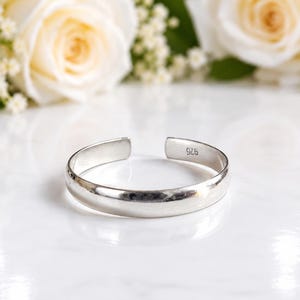 Könnte beinhalten: Ein polierter Silberarmreif mit glatter, abgerundeter Oberfläche und offenem Design. Auf der Innenseite des Bandes ist deutlich der Stempel 925 eingraviert, der die Metallzusammensetzung angibt. Das Armband hat eine schlichte, klare Optik.