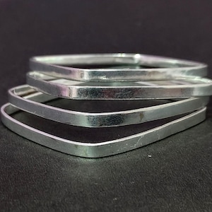 Op de afbeelding: Vier zilveren vierkante armbanden gestapeld op een donkere ondergrond. De armbanden hebben een platte, gepolijste afwerking en een afgeronde binnenrand. De armbanden zijn gemaakt van glanzend metaal.