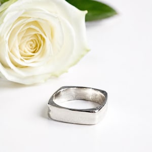 Könnte beinhalten: Ein polierter silberfarbener Ring mit einem markanten quadratischen Außenband und einer kreisförmigen Innenöffnung. Die Metalloberfläche ist glatt und reflektierend und zeigt ein klares, geometrisches Design, das flach aufliegt.