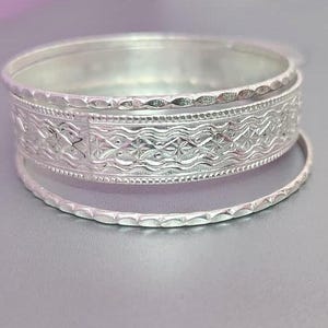 Op de afbeelding: Een stapel zilveren armbanden. De grootste armband heeft een ingewikkeld reliëfontwerp met bloemen- en golfpatronen. Twee kleinere, dunnere armbanden zijn ook zichtbaar, waardoor een gelaagde look ontstaat. De sieraden worden gepresenteerd op een lichtgrijze ondergrond.